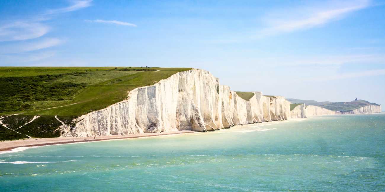 Discover the untamed beauty of the Seven Sisters Cliffs along the Sussex shoreline in 2026 High-angle panoramic view of the massive white chalk Seven Sisters Cliffs sweeping along the bright turquoise waters under a blue sky