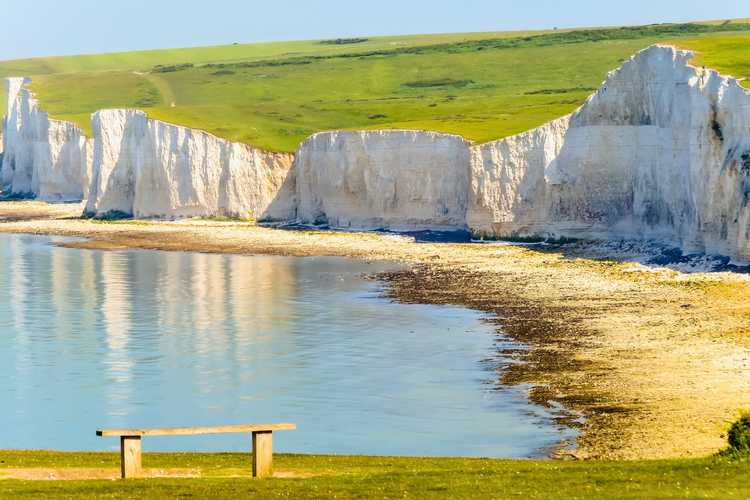 A peaceful clifftop viewpoint looking along the historic Seven Sisters Cliffs in Sussex in 2026 View from a green grassy hill with a rustic wooden bench looking toward the immense white chalk walls of the Seven Sisters Cliffs and calm sea